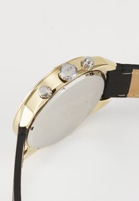 Reloj de acero inoxidable dorado con correa de cuero negro, que presenta una caja redonda, pulsadores plateados y un dorso liso y pulido.