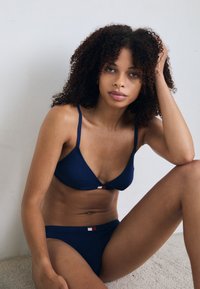 Mørkeblå bikini sett i et glatt stoff med tynne stropper, med en liten logo aksent i midten, iført i en avslappet positur.