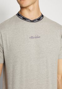 Mężczyzna w jasnoszarej koszulce ellesse z czarnym kołnierzykiem z logo, widoczny od szyi do środka torsu.