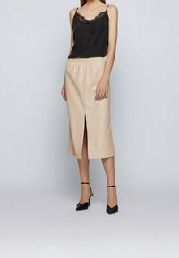 Camisole hitam dengan renda di tepi dan tali tipis, dipadukan dengan rok faux leather berwarna beige sepanjang midi yang memiliki belahan di bagian depan. Sandal hak hitam.