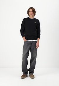 Zwarte sweatshirt met een witte kraagdetail, voorzien van een klein geel logo op de borst, gecombineerd met wijde donkergrijze jeans en bruine schoenen.