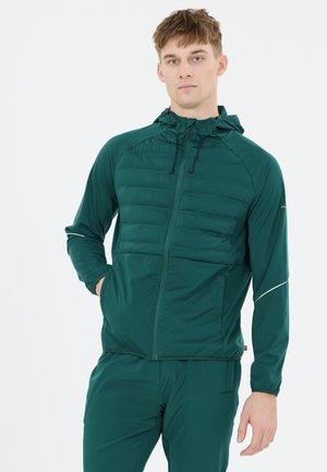Mann, der eine grüne Kapuzen-Trainingsjacke und passende Hose trägt, steht mit einer Hand in der Tasche und blickt nach vorne vor einem schlichten Hintergrund.