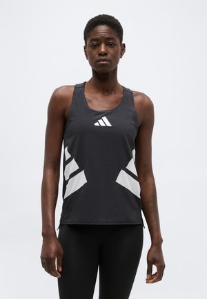 Atletische vrouw die een zwarte mouwloze Adidas tanktop met witte logo's draagt, staand tegen een effen lichte achtergrond.