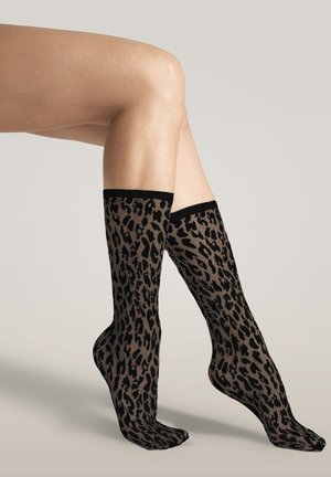 Benen gekruist bij de knie, met doorschijnende kniekousen met een zwart-bruine luipaardprint tegen een neutrale achtergrond.