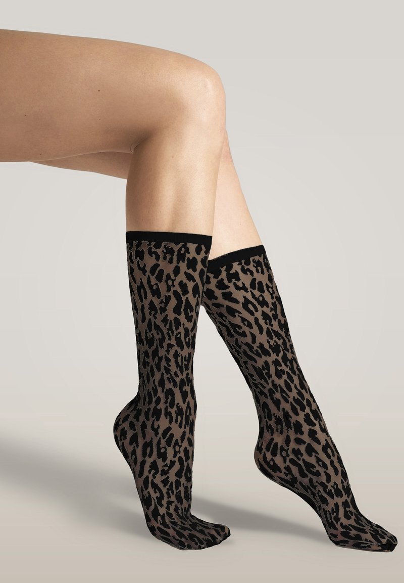 Gambe incrociate al ginocchio, con calze trasparenti fino al ginocchio a motivo leopardato nero e marrone su uno sfondo neutro.