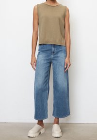 Person wearing sleeveless beige knit top, wide-leg blue jeans, and chunky off-white slip-on shoes, standing against white wall.