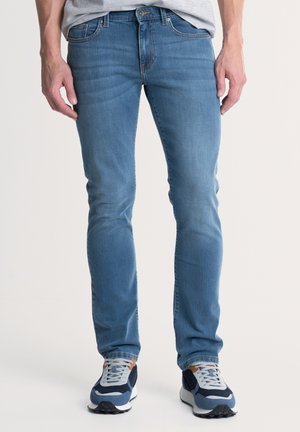 Mėlynos džinsinės kelnės su slim fit fasonu, turinčios penkių kišenių dizainą ir subtilaus blukimo efektą. Derinamos su pilkomis, mėlynomis ir baltomis sportinėmis bateliais.