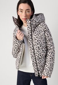 Eine junge Frau präsentiert eine leopardgemusterte Pufferjacke mit Kapuze, hält den Reißverschluss und trägt ein helles Oberteil sowie dunkle Jeans.