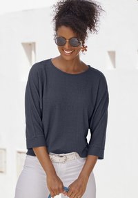 Navyfarbener, strukturierten Langarm-Top mit weitem Ausschnitt, kombiniert mit weißen Hosen und einem hellen Gürtel. Modell hält ein blaues Accessoire.