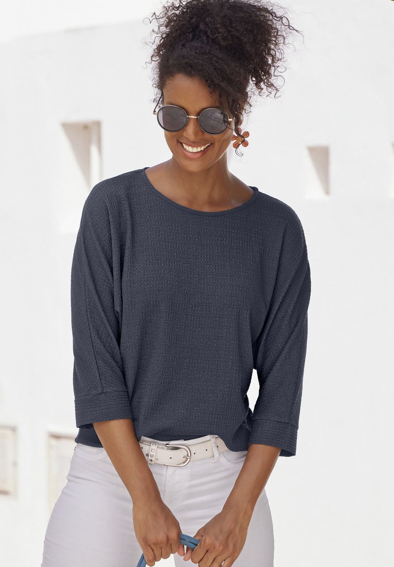 Navyfarbener, strukturierten Langarm-Top mit weitem Ausschnitt, kombiniert mit weißen Hosen und einem hellen Gürtel. Modell hält ein blaues Accessoire.