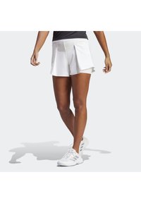 Pantalones cortos deportivos blancos con diseño de pliegues, que presentan una textura suave y pantalones cortos interiores integrados, combinados con zapatos deportivos blancos.