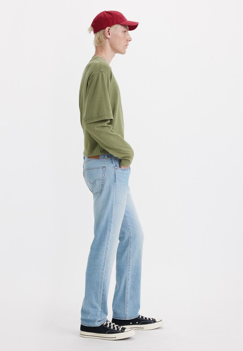 Chemise verte à manches longues, jean bleu clair, baskets noires avec semelles blanches et casquette rouge. Le modèle se tient de côté devant un fond blanc.