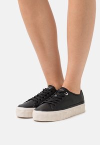 Černé tenisky s texturovaným materiálem, zaobleným tvarem a bílou vzorovanou podrážkou. Disponují šněrovacím designem a kovovými očky.