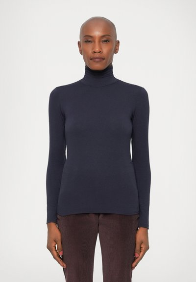 Maglione a collo alto di colore navy con maniche lunghe, realizzato in tessuto morbido, con un design aderente e costruzione senza cuciture sulle spalle, abbinato a pantaloni scuri.