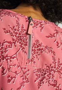Gros plan d'une robe fleurie rose avec une fermeture éclair argentée visible et un tire-zip rose au niveau de l'encolure arrière.