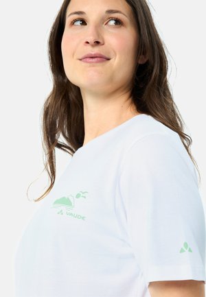 Femme aux longs cheveux bruns portant un t-shirt blanc Vaude avec un logo vert de montagne et d'oiseau sur la poitrine et la manche, regardant vers le haut.