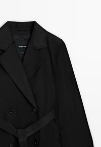 Trench-coat noir croisé avec col à revers et ceinture nouée à la taille, posé à plat sur fond blanc.