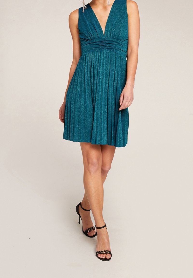 Femme portant une robe plissée sans manches de couleur sarcelle avec un décolleté en V profond et des sandales noires à talons hauts avec brides à la cheville, debout sur un fond clair.