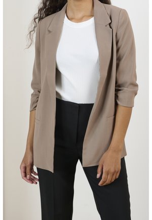 Kobieta ubrana w beżową marynarkę z podwiniętymi rękawami, biały prążkowany top i czarne spodnie, stoi na tle jednolitego jasnego tła.