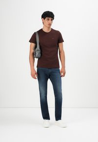 Junger Mann steht auf einem weißen Hintergrund und trägt ein weinrotes T-Shirt, blaue Jeans, weiße Sneakers und eine graue Umhängetasche.