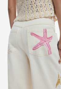 Krémové nohavice s dvoma zadnými vreckami, ktoré sú ozdobené vyšívanými ruzovými hviezdicami a mušľami. Materiál je hladký denim so dekoratívnym šitím.