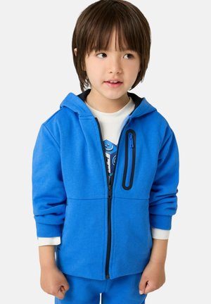 Felpa zip-up blu in tessuto morbido, con una tasca con zip nera e polsini a coste, abbinata a una maglietta bianca con grafica sotto.