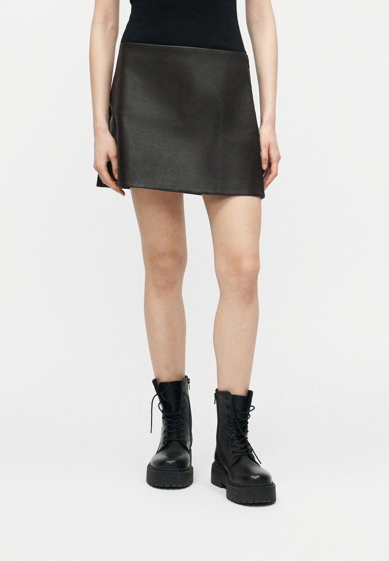 Άτομο που φοράει κοντή μαύρη δερμάτινη φούστα με μαύρα ψηλοτάκουνα μποτάκια με κορδόνια, στέκεται μπροστά σε απλό φωτεινό φόντο.