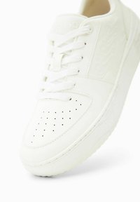 Baskets blanches avec une boîte à orteils perforée, tige en cuir lisse, lacets plats et semelle en caoutchouc texturée. Marque discrète sur la languette.