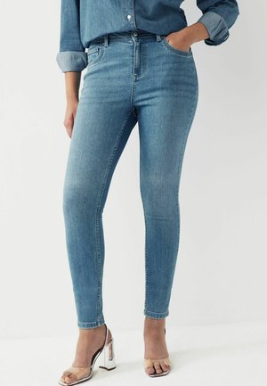 Jeans en denim slim fit bleu, avec une taille haute, poches avant bien définies et légère décoloration. Associés à des sandales à talons transparents.