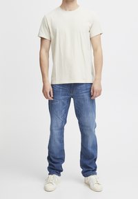 Homme portant un t-shirt à manches courtes crème, un jean bleu et des baskets blanches, se tenant devant un fond blanc uni.