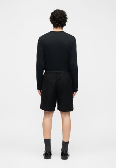 Άτομο με σγουρά μαλλιά στέκεται πλάτη, φοράει μαύρο μακρυμάνικο πουκάμισο, μαύρα σορτς με σχέδια, σκουρόχρωμες κάλτσες και μαύρα παπούτσια.