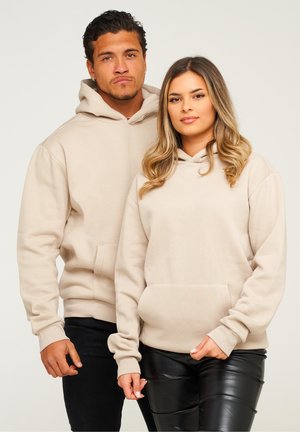 Beige hoodies with kangaroo pockets, long sleeves, and relaxed fit. The male model wears black jeans, while the female model wears black leather pants.