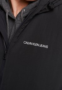 Svart ytterjacka med dragkedja framtill, lager på lager över en grå huvtröja. Har vit "CALVIN KLEIN JEANS"-logotyp på bröstet. Mjuk yta.