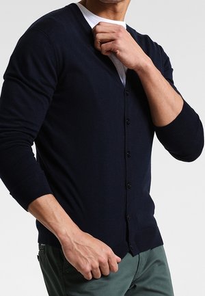 Uomo che indossa un cardigan blu navy scuro con bottoni sopra una camicia bianca, mentre sistema il colletto con la mano destra, in piedi davanti a uno sfondo neutro.