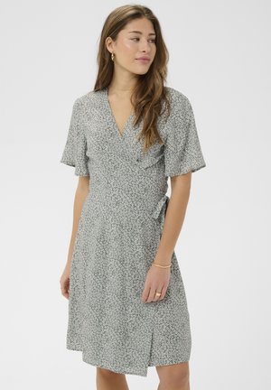 Mlada ženska v sivi kratkoročni preobleki z rožnatim vzorcem in zavezovanjem na strani, ki gleda vstran na enostavni ozadju.