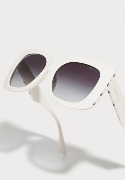 Lunettes de soleil blanches oversize carrées avec verres dégradés foncés et clous décoratifs sur les branches sur fond clair.