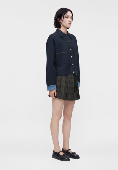 Νεαρή γυναίκα που φοράει ένα σκούρο σακάκι με κουμπιά και μανσέτες από τζιν, μια πλισέ σκουρόχρωμη φούστα και μαύρα παπούτσια Mary Jane, στέκεται μπροστά σε απλό φόντο.