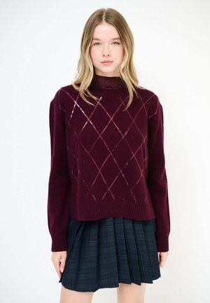 Giovane donna che indossa un maglione a rombi bordeaux scuro sopra un dolcevita e una gonna plissettata verde scuro, in piedi su uno sfondo bianco semplice.