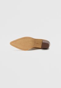 Pantof suede maro deschis cu toc înalt, vârf ascuțit și toc neted de culoare brună. Dispune de o talpă cusută cu un model texturat pe margine.
