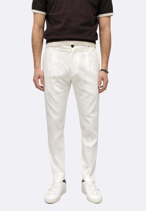 Uomo con pantaloni bianchi plissettati, scarpe da ginnastica bianche e camicia scura a maniche corte, in piedi con le mani rilassate lungo i fianchi.