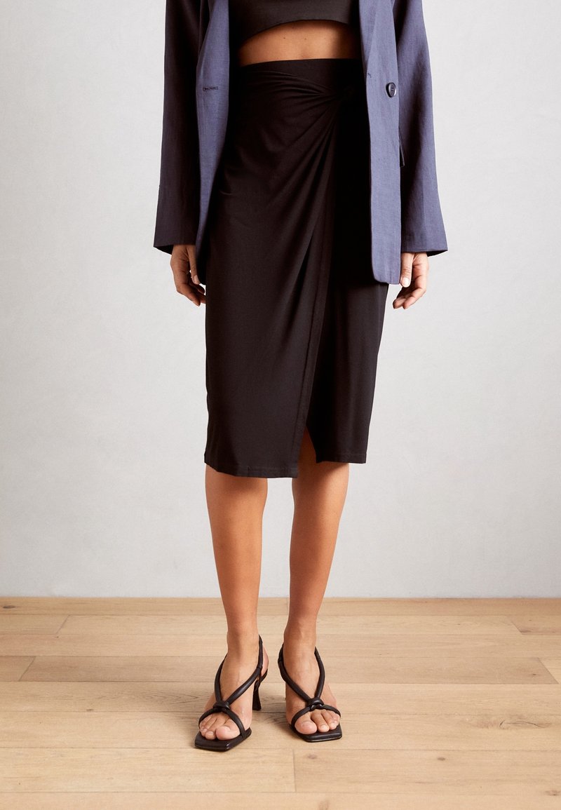 Femme portant une jupe portefeuille noire, des sandales à talons noires à lanières, un haut court noir et un blazer bleu, debout sur un sol en bois clair.