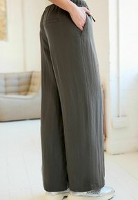 Pantaloni a gamba larga in un tessuto verde oliva tenue, dotati di una cintura elastica e tasche laterali, abbinati a sneaker strutturate.
