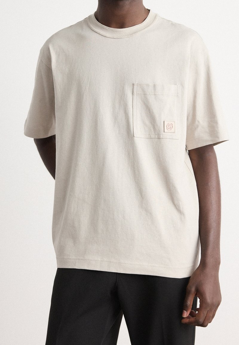 Beige bomulls t-shirt med en avslappnad passform, rund halsringning, korta ärmar och en enda bröstficka med en liten logotypapplikation.