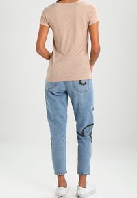 Beige kortærmet top, lyse jeans med sorte broderede detaljer, og hvide sneakers, set fra ryggen mod en ensfarvet baggrund.