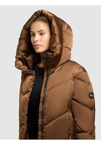 Veste matelassée marron avec une grande capuche, fermeture éclair à l'avant, boutons-pression et texture matelassée. Comprend un patch logo sur la manche gauche.