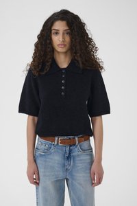 Frau mit lockigem Haar trägt einen schwarzen, kurzärmligen, geknöpften Pullover, hellblaue Jeans und einen braunen Gürtel und steht vor einem schlichten Hintergrund.