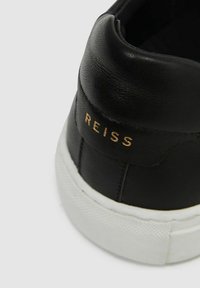 Schwarze Leder-Sneaker mit einer weißen Gummisohle und goldener "REISS"-Branding am Fersenbereich. Glatte Textur mit einem flachen Design.