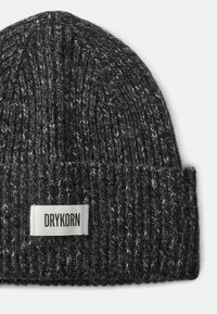 Pletená černá čepice s ohrnutým lemem, která má světle šedou nášivku s nápisem "DRYKORN." Textura je silná a pohodlná, se smíšenými barvami příze.