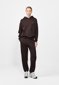 Moteris, vilkinti tamsiai rudą džemperį ir derančius sportinius kelnes, su baltais sportbačiais, stovi prieš paprastą šviesią fono.