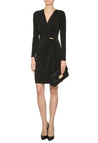 Femme portant une robe portefeuille noire à manches longues avec une boucle dorée, tenant une pochette noire et portant des talons noirs pointus.